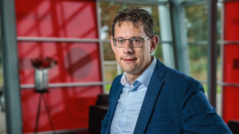 Man met bril in blauw colbert poseert binnen voor rood-witte wand