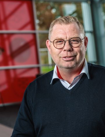 Man met bril en blond haar in donkerblauwe trui voor rood-witte wand