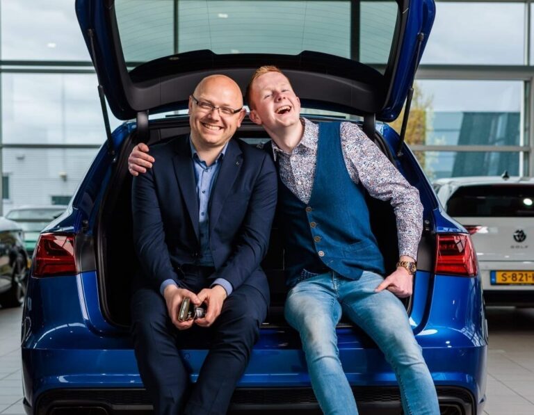 Twee lachende mannen zitten in de open kofferbak van een blauwe auto