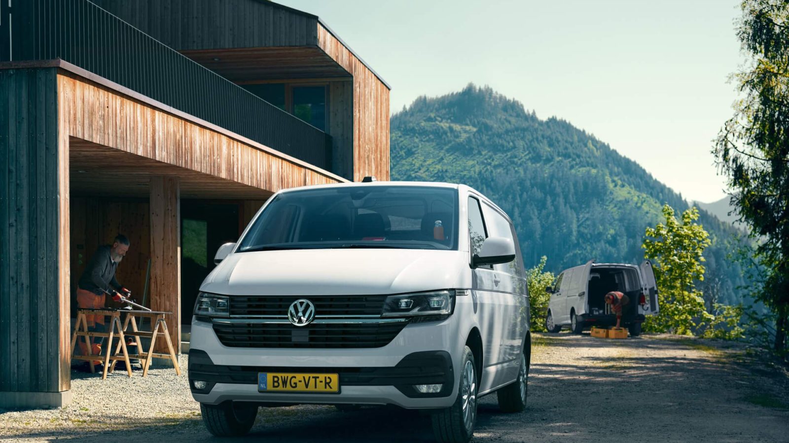Witte bestelbus voor een houten gebouw in bergachtig landschap, met twee werkende mensen