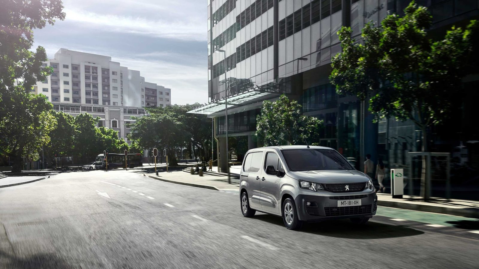 Zilveren Peugeot bestelwagen rijdt over een brede stadsstraat langs moderne glazen gebouwen