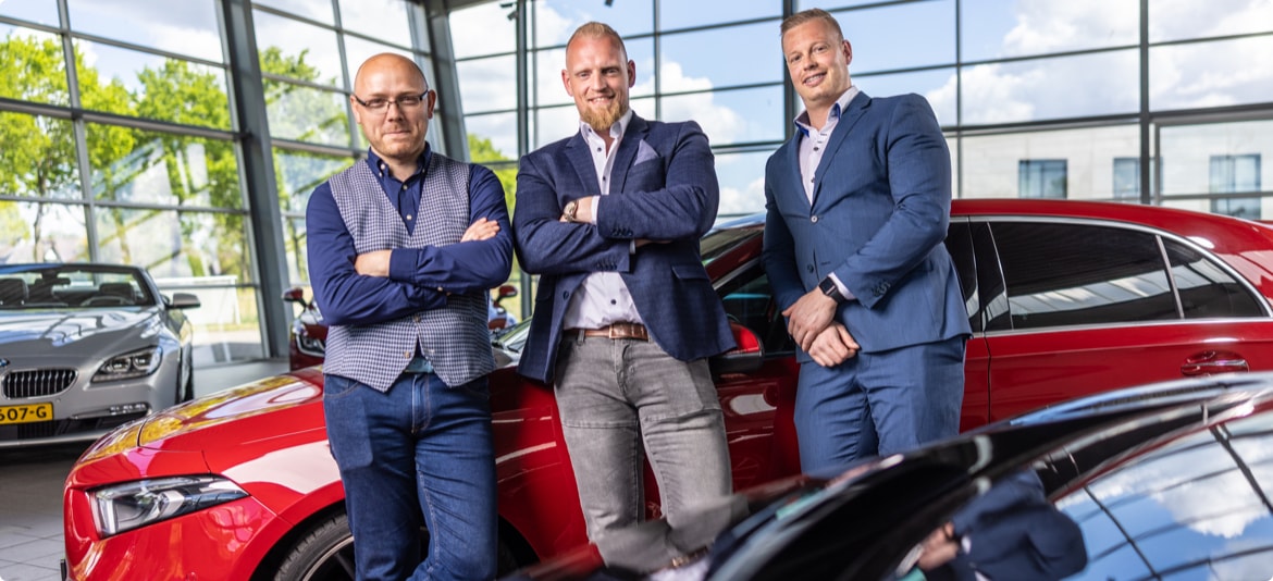 Drie mannen staan met gekruiste armen bij een rode auto in een showroom met grote ramen