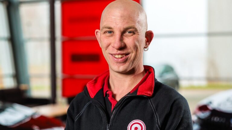 Glimlachende kale man met oorbel in zwarte trui met rood logo, op de voorgrond binnen