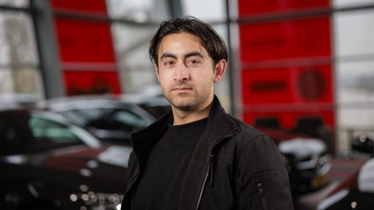 Man met donker haar in zwarte jas poseert binnen bij auto's