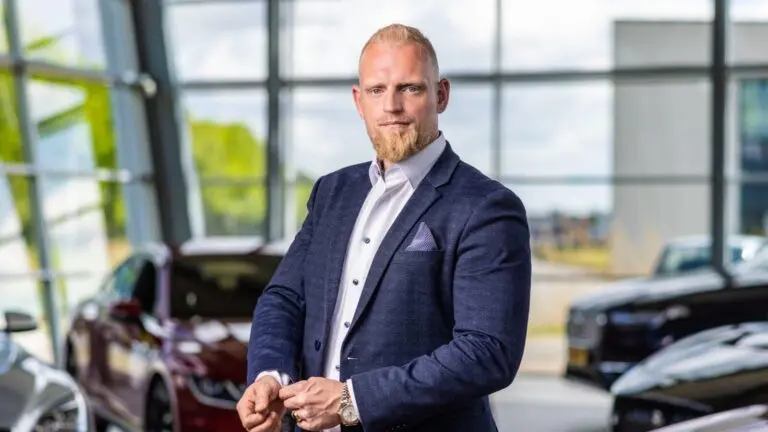 Man in pak staat in een showroom met auto's op de achtergrond