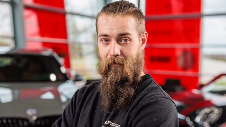Man met volle baard en opgestoken haar poseert met gekruiste armen voor een auto