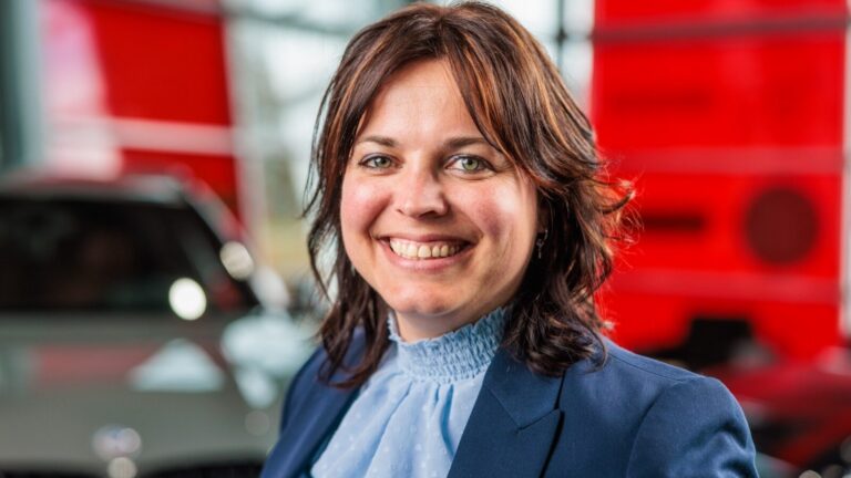 Vrouw met bruin haar en blauwe blouse glimlacht in een moderne omgeving met rode achtergrond