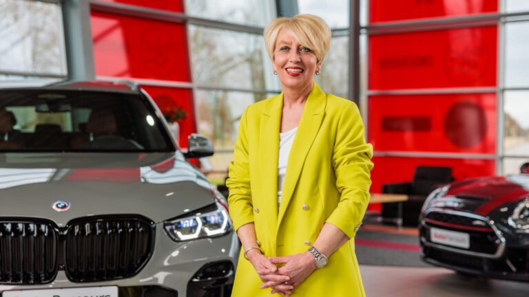 Vrouw met blond kort haar in gele blazer staat binnen naast grijze BMW