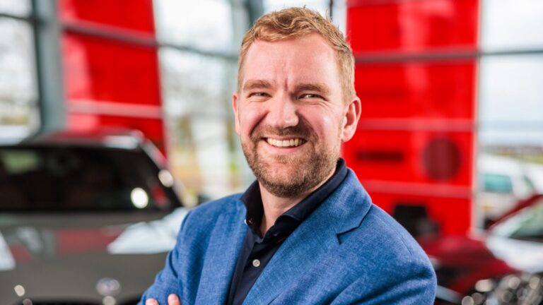 Man met baard in blauw colbert glimlacht in een showroom met rode panelen op de achtergrond