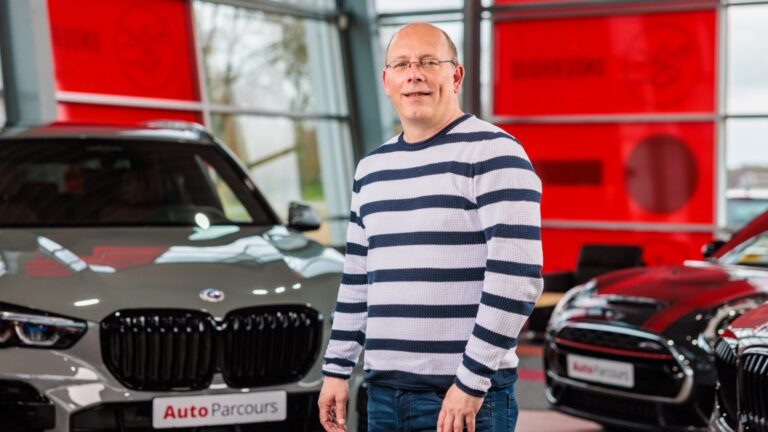 Man met bril en gestreepte trui staat binnen tussen twee auto's